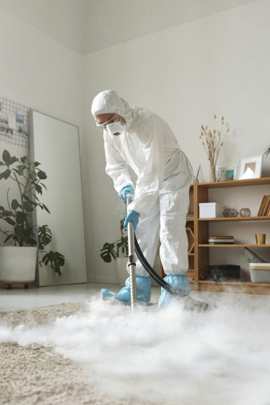 Person in protective gear using a vacuum cleaner on a carpet with visible clouds of cleaning foam.
